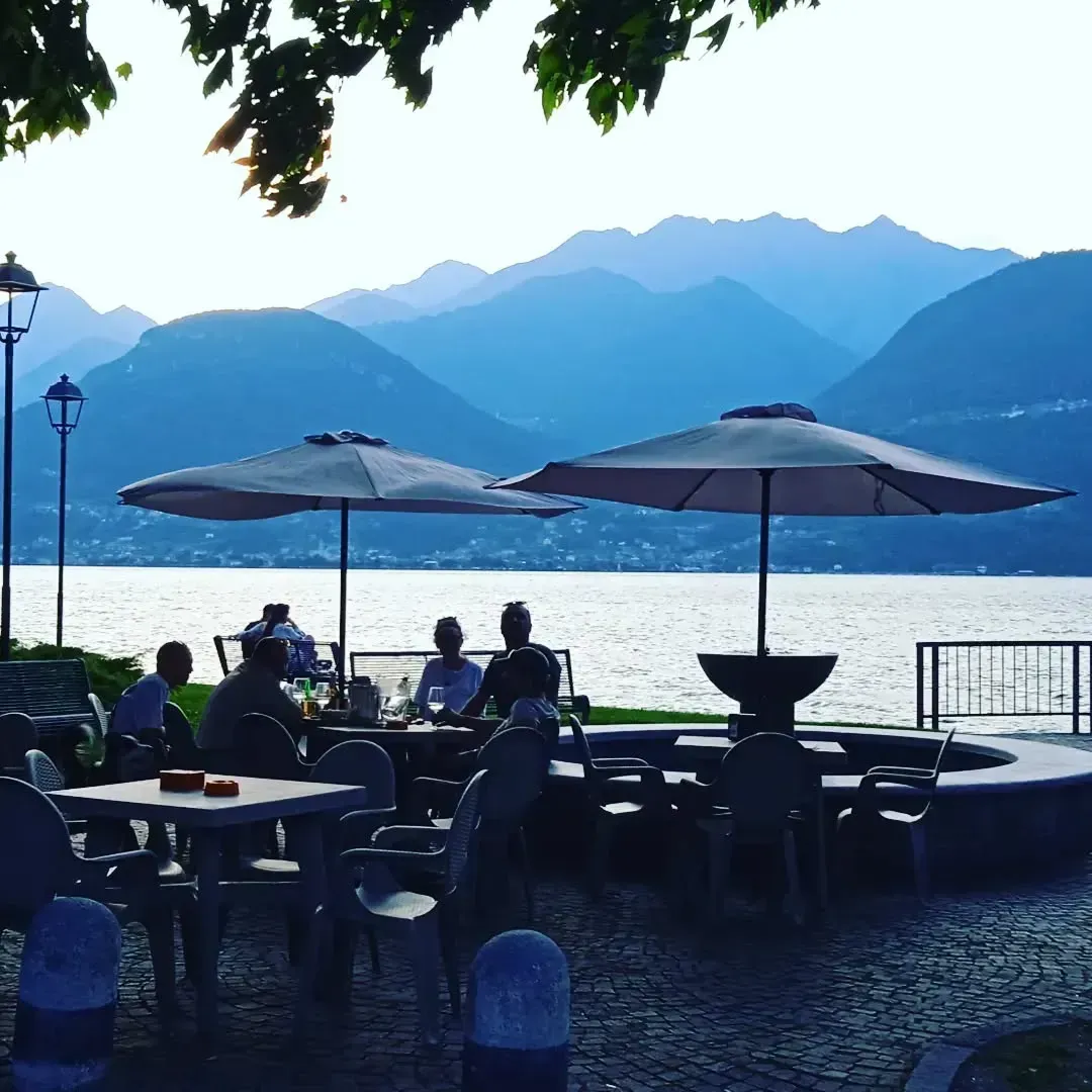 Bar all'aperto con gente seduta ai tavoli, in riva al lago con le montagne sullo sfondo.