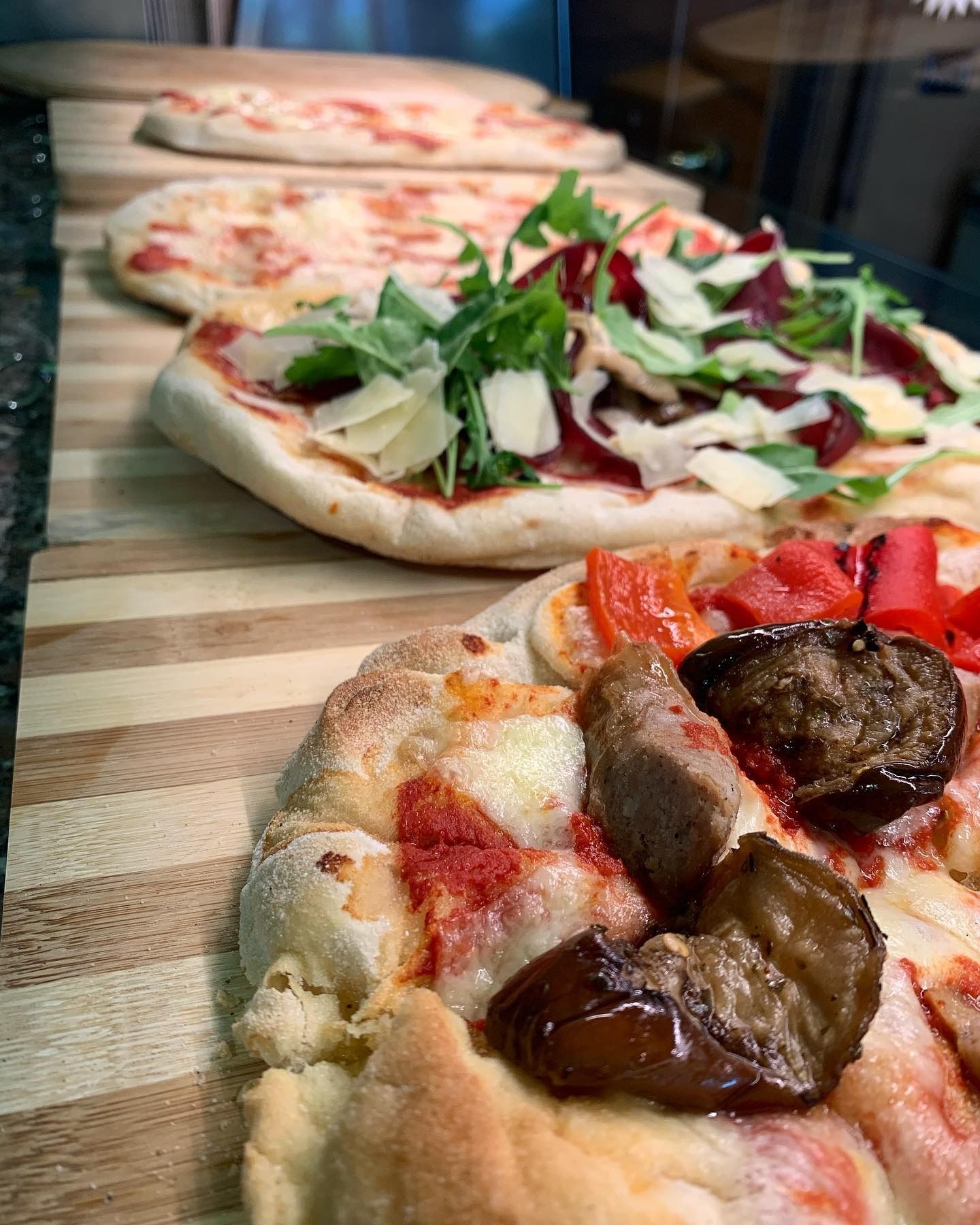 Diverse pizze su un tagliere di legno a strisce, condimenti visibili: rucola, formaggio, salsiccia, melanzane, pomodori.