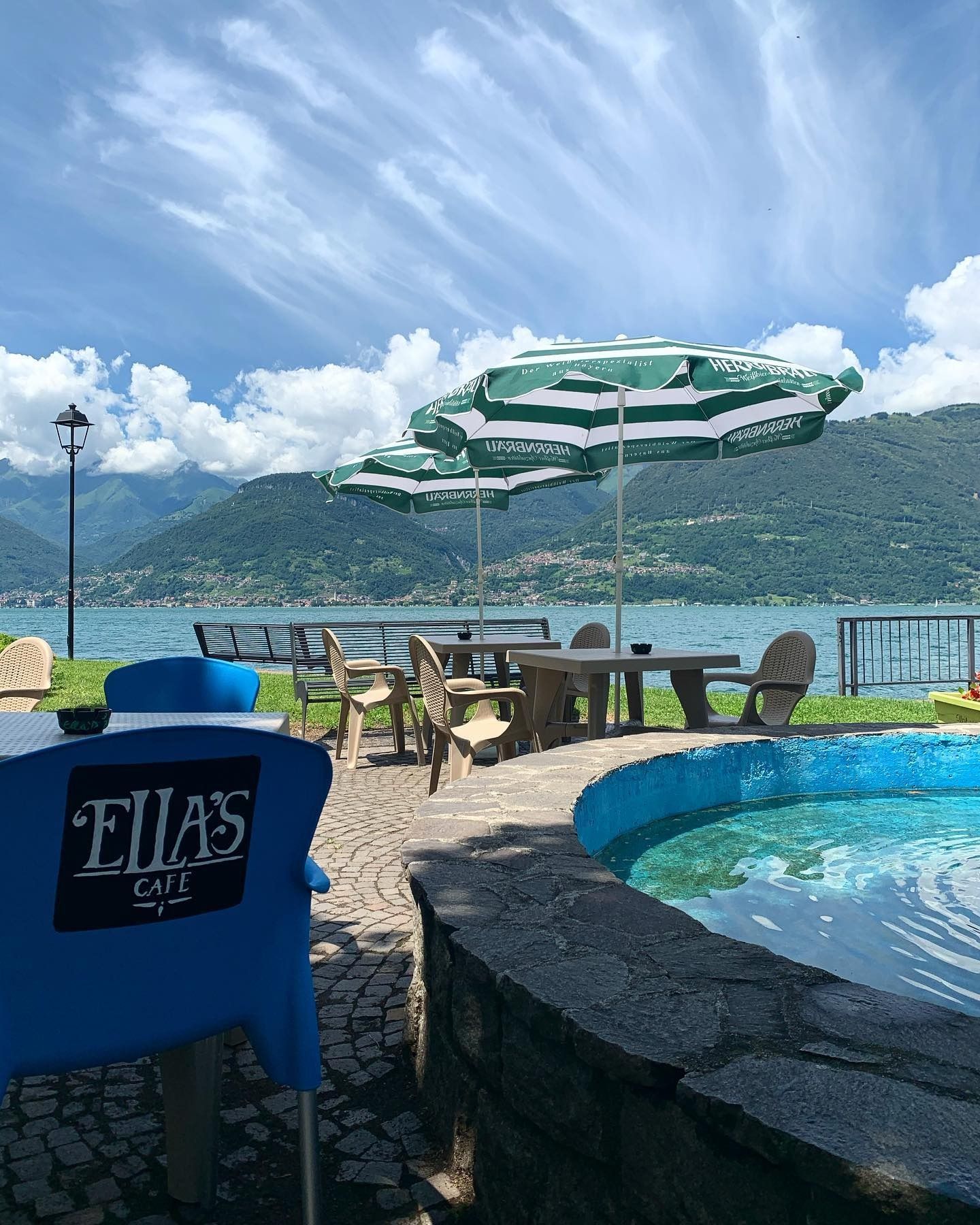 Bar all'aperto con vista sul lago, ombrelloni verdi, acqua azzurra, giornata di sole.