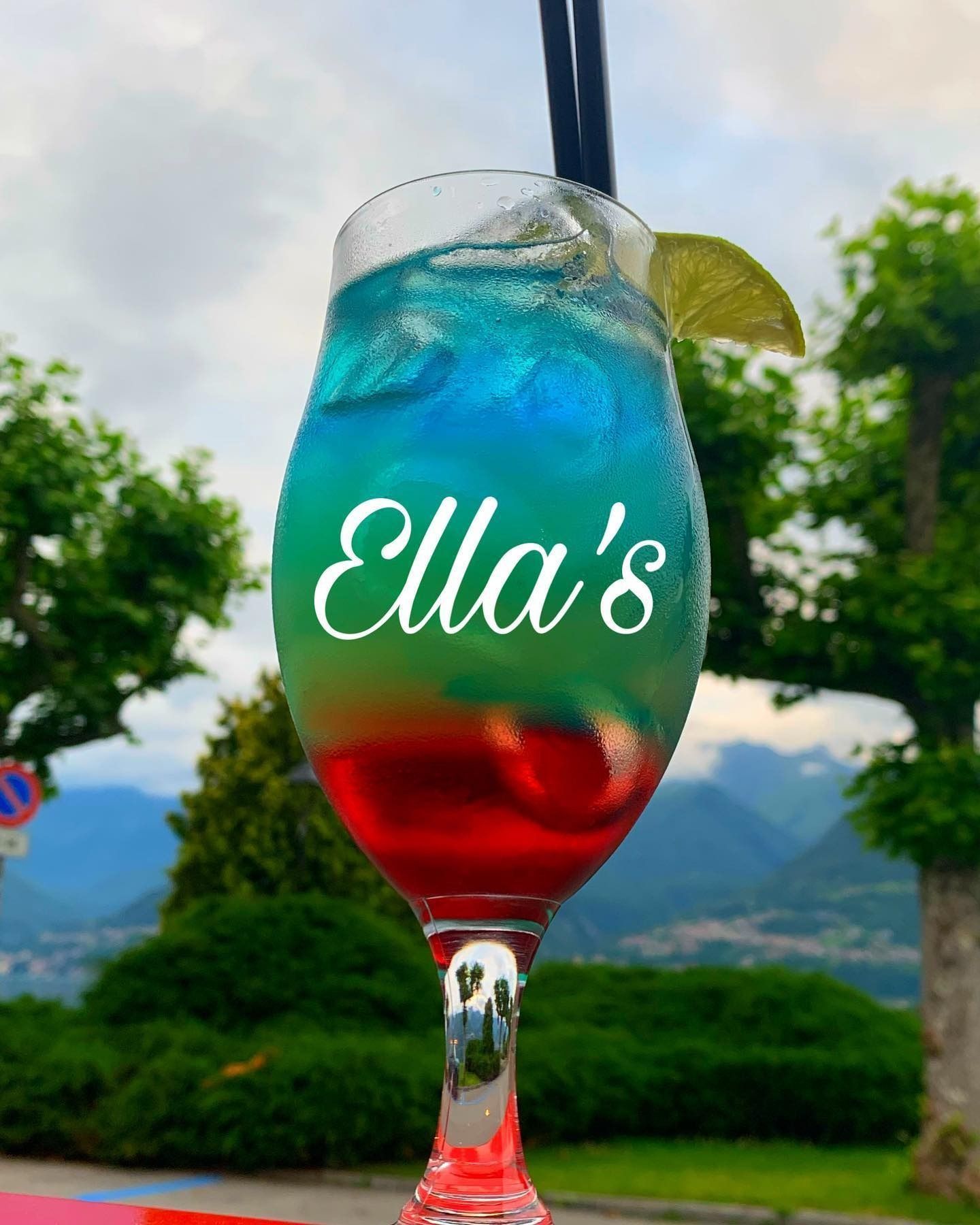 Cocktail colorato a strati in un bicchiere alto, logo di Ella sul bicchiere, spicchio di lime, due cannucce, ambiente esterno.