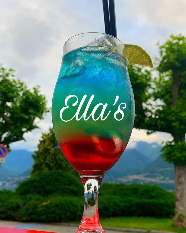 Cocktail colorato a strati in un bicchiere alto, logo di Ella sul bicchiere, spicchio di lime, due cannucce, ambiente esterno.