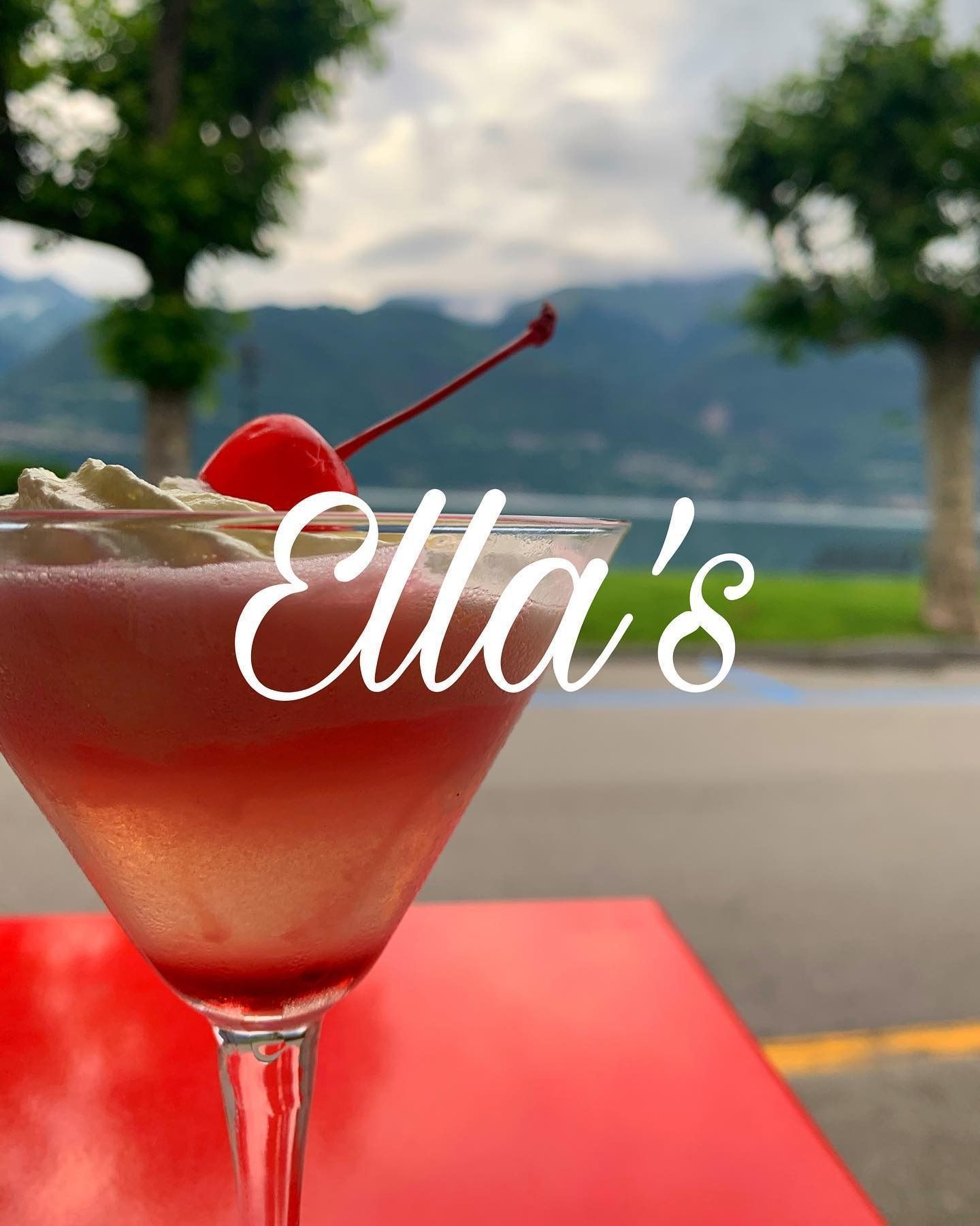 Cocktail in un bicchiere da martini con ciliegia, panna montata e scritta "Ella's" sovrapposta. Tavolo rosso, sfondo panoramico.