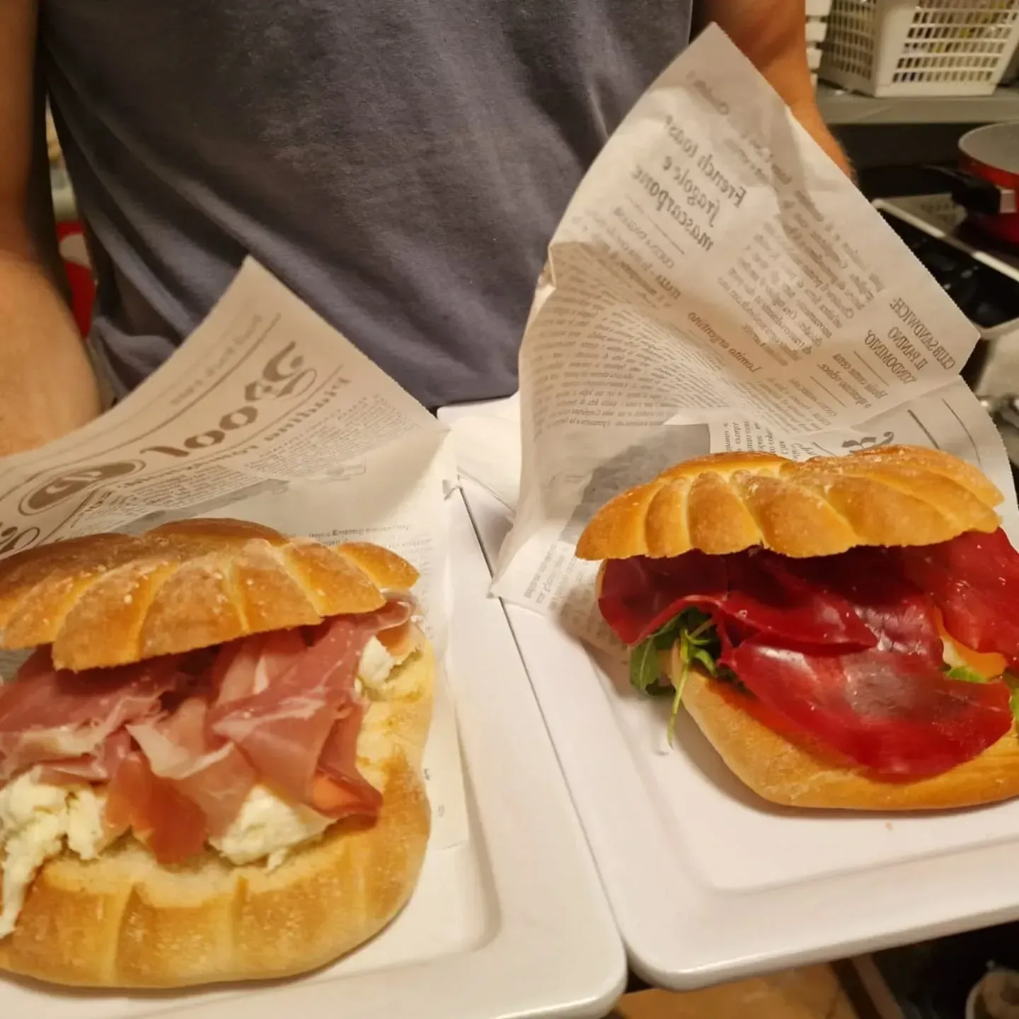 Due panini su piatti bianchi, avvolti nella carta: uno con prosciutto e formaggio, uno con carne rossa.