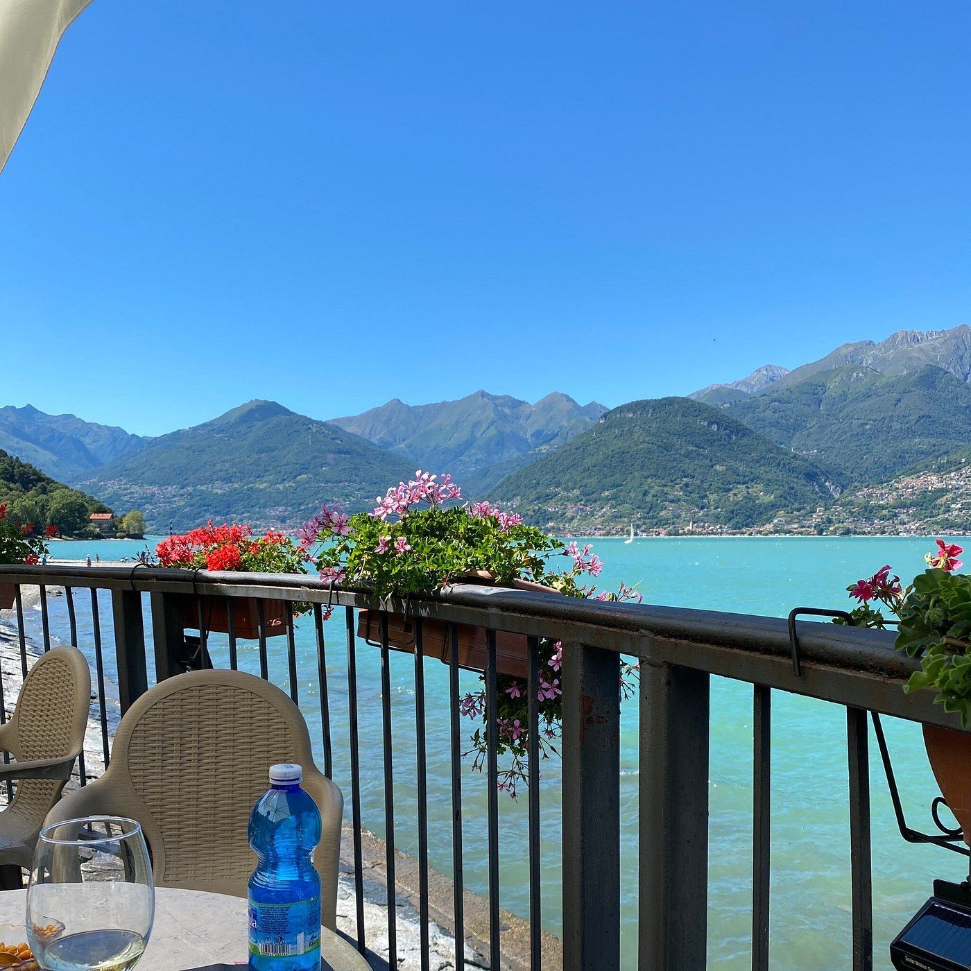 Balcone con vista sul lago turchese e sulle montagne, con fiori, un tavolo e una bottiglia d'acqua.