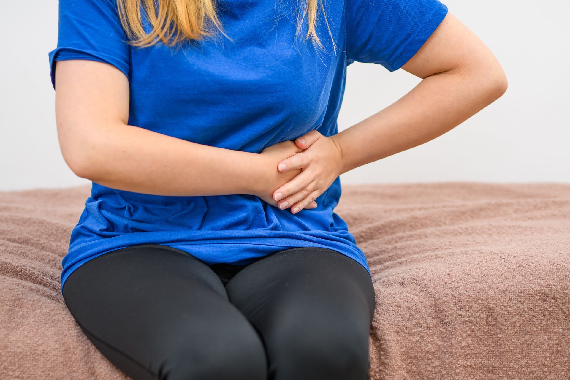 A woman in a blue shirt is sitting on a bed holding her stomach.