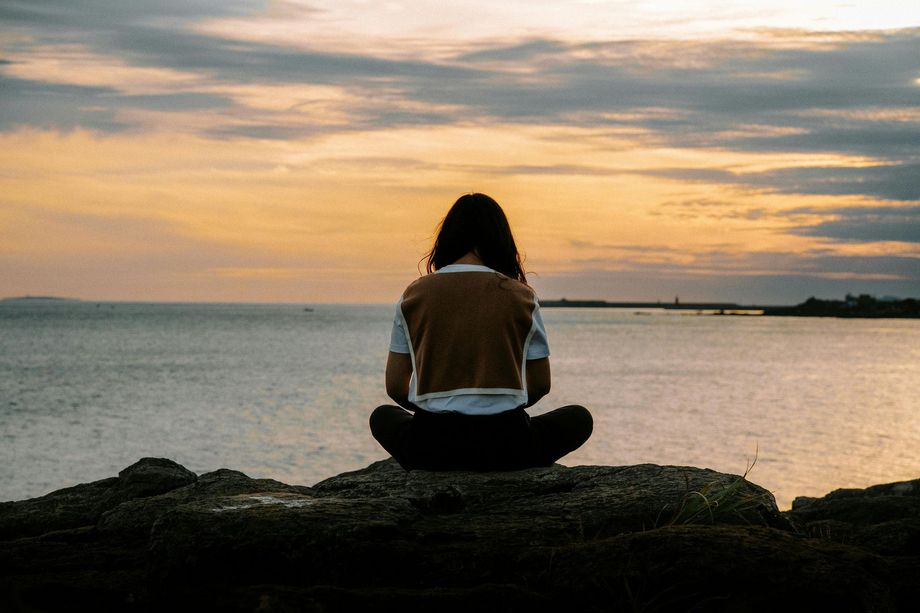 A woman is sitting on a rock near the ocean at sunset.