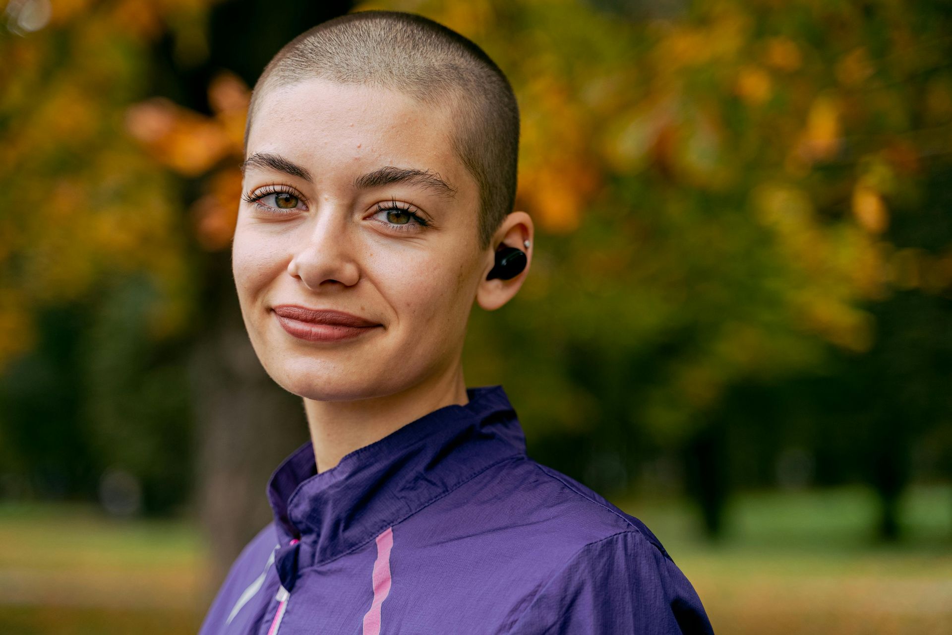 A woman with a shaved head is wearing headphones and a purple jacket.