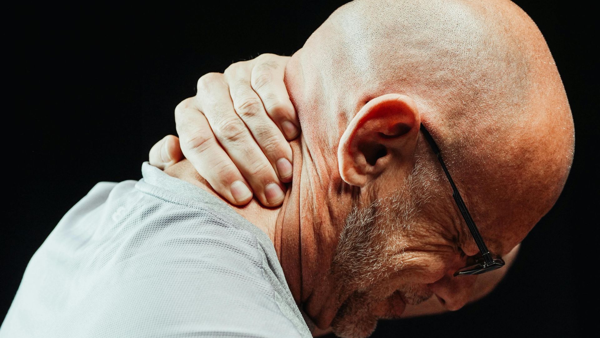 A bald man with glasses is holding his neck in pain.