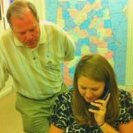 Man leans over woman talking on phone; map on wall in background.