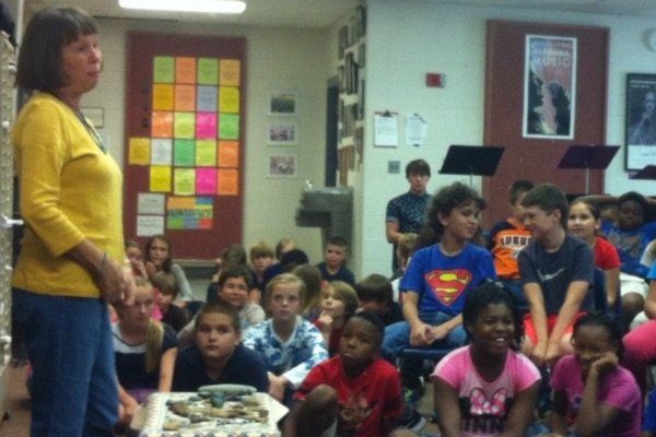 Woman speaking to group of children in classroom, colorful notes on the wall, children looking at the speaker.