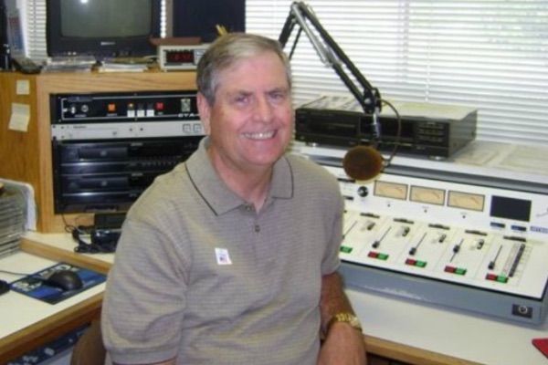 Man smiling, seated at radio station equipment. Beige shirt, microphone, control board, and dials visible.