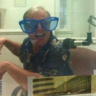 Man wearing oversized blue glasses, smiling, in a radio booth, leaning toward microphone.