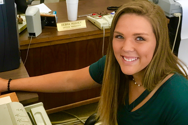 Smiling person at a desk. Wearing green shirt, pearl necklace. Blonde hair, tan complexion.