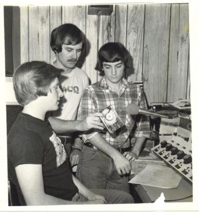 Three young men in a studio examining a record. One holds it up, others observe.  Control board visible.