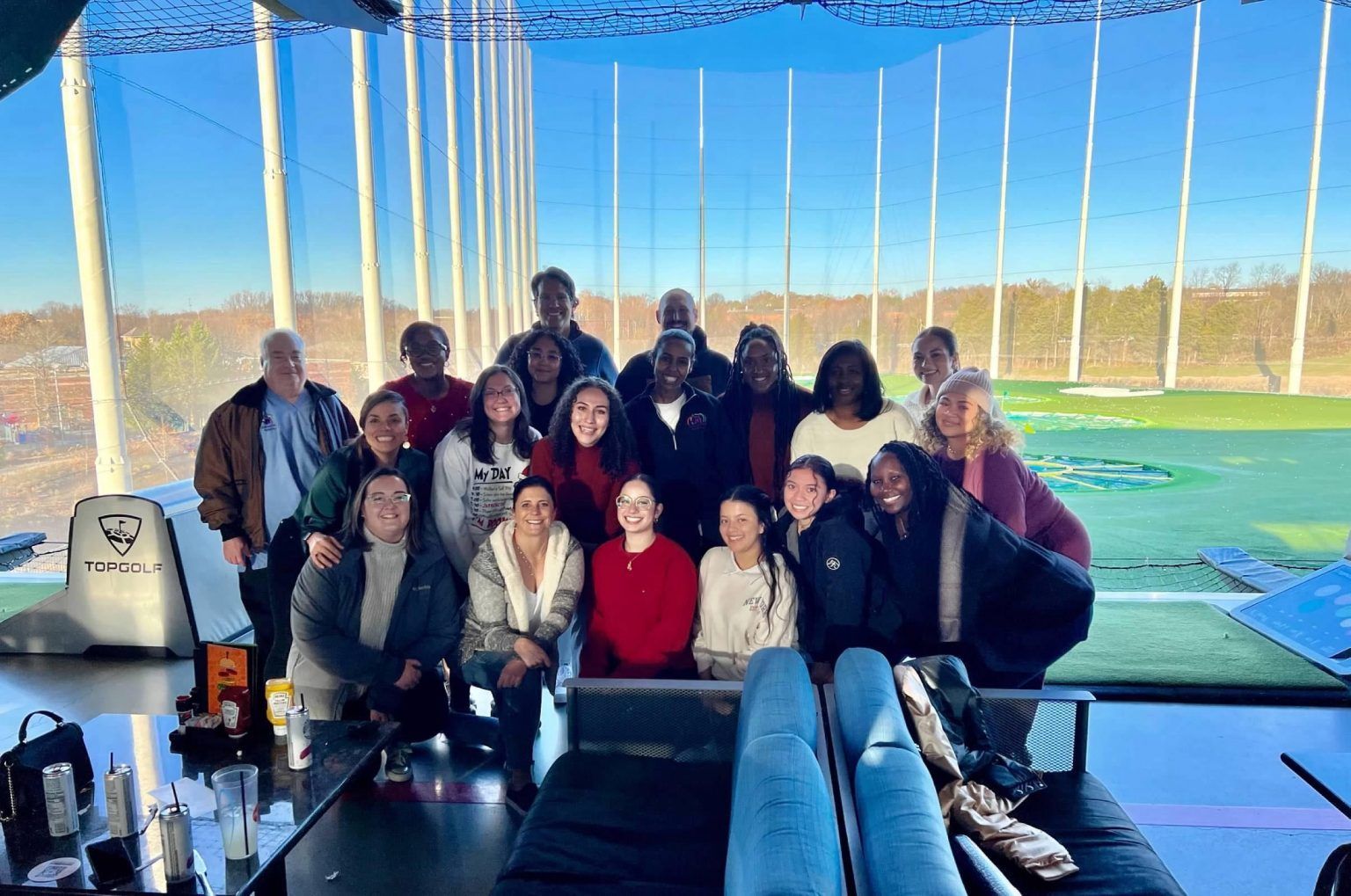 Group of people smiling at a Topgolf location. Sunny day with people in casual winter clothing.