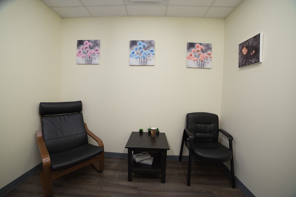 A counseling room with beige walls, three floral paintings, and two black chairs facing a small table.