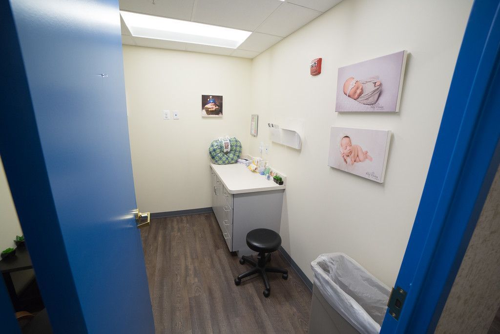 A doctor's examination room with light blue door frame. Room features a cabinet, stool, and baby-themed artwork.
