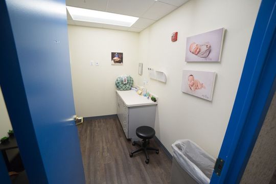 A doctor's examination room with light blue door frame. Room features a cabinet, stool, and baby-themed artwork.