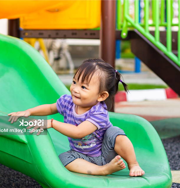 A young Asian girl smiles while sitting on a green playground slide. She wears a purple shirt and gray pants.