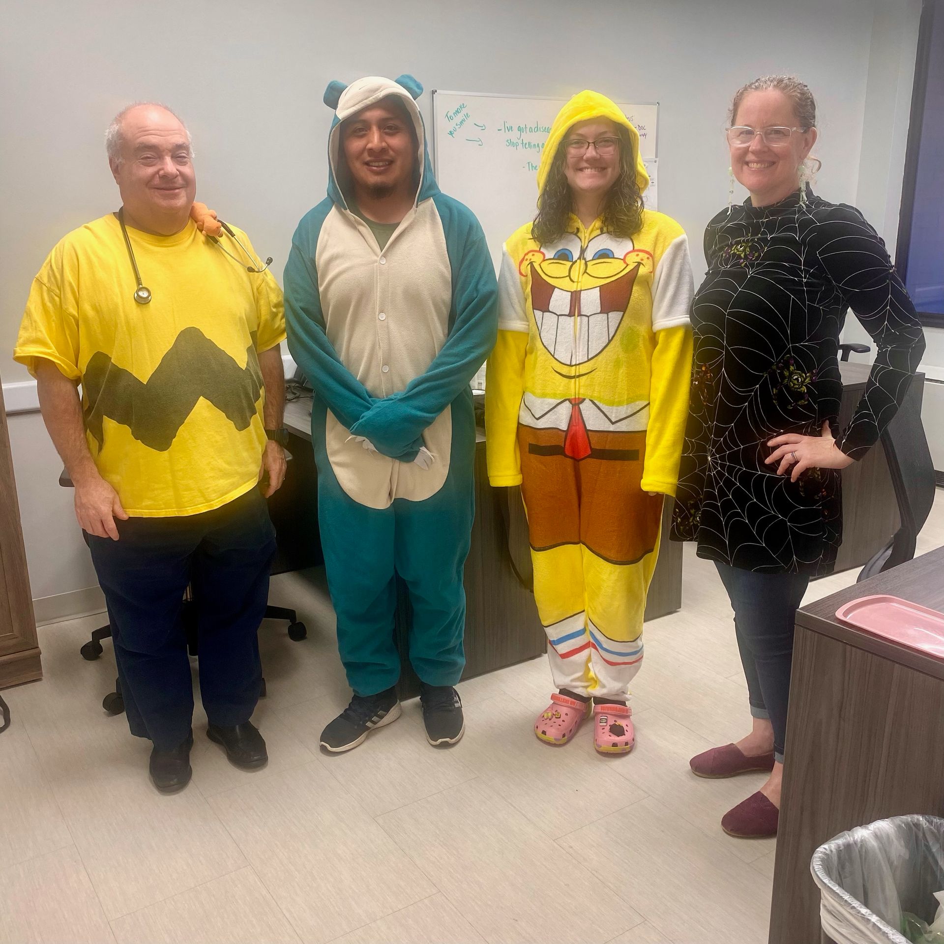 Four people in a room, dressed in costumes: Charlie Brown, a blue bear, Spongebob Squarepants, and a woman in a black dress.
