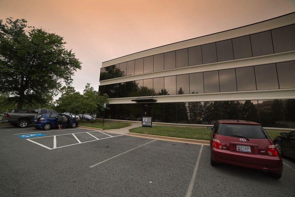 Office building with a glass facade and a parking lot. Red car parked on the right, blue truck in the left, and a wheelchair parking spot.