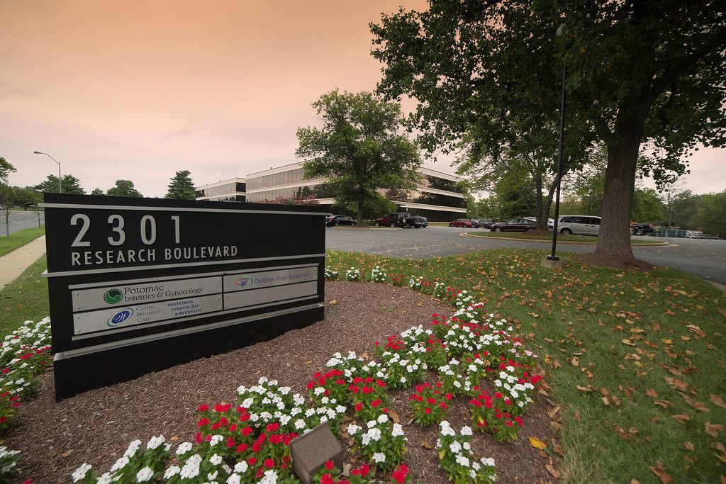 Sign for 2301 Research Blvd, with red and white flowers in front. Building and trees are in the background, under a cloudy sky.