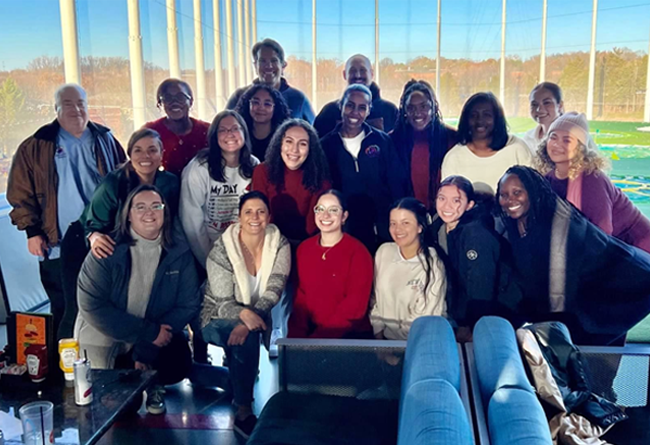 Group photo of diverse people smiling, inside a golf driving range, with windows overlooking a green field and trees.