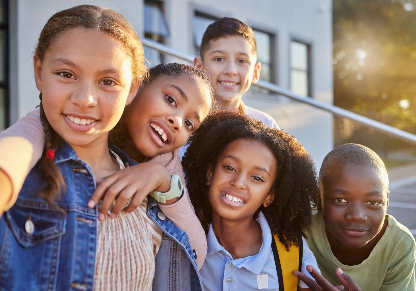 Group of smiling children posing outdoors. Several have their arms around each other. Sunny, school setting.