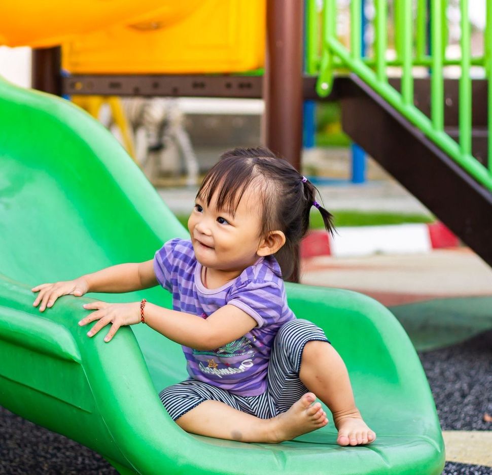 Young child with pigtails smiles while sitting on a green playground slide. They are wearing a purple striped shirt and black and white striped pants.