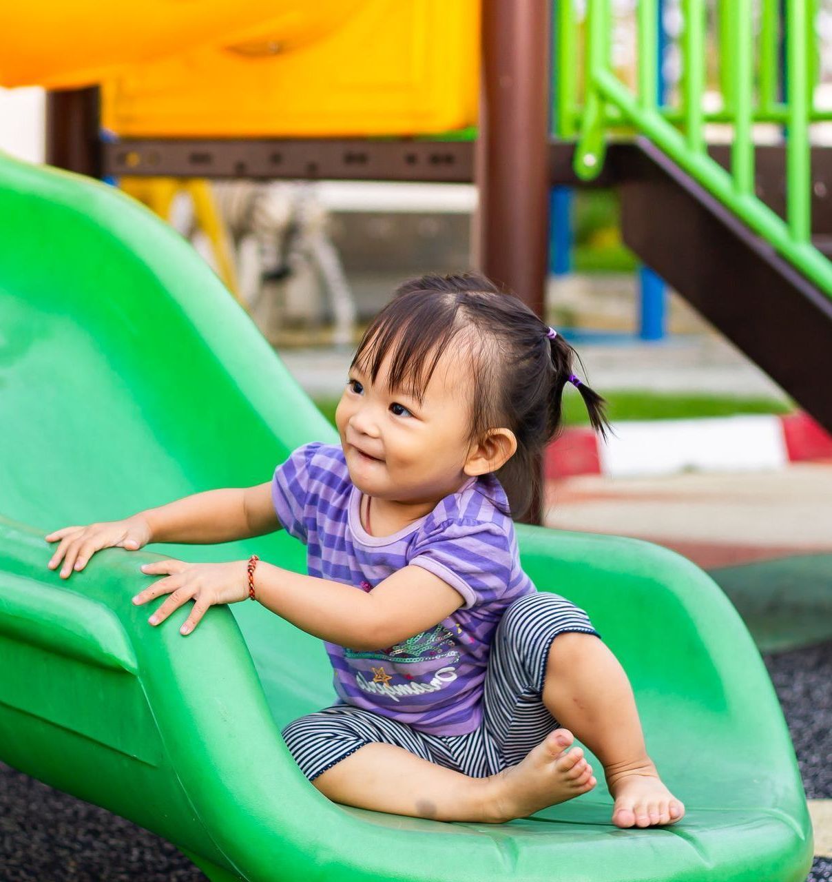 Toddler sitting on a green playground slide, smiling and holding the edge