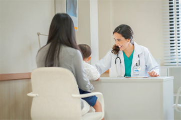 A doctor smiles while speaking to a mother holding a child at a desk in a medical office.