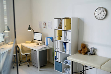 A doctor's office interior with a desk, shelves, examination table, and clock. White walls, organized files, and a teddy bear are visible.