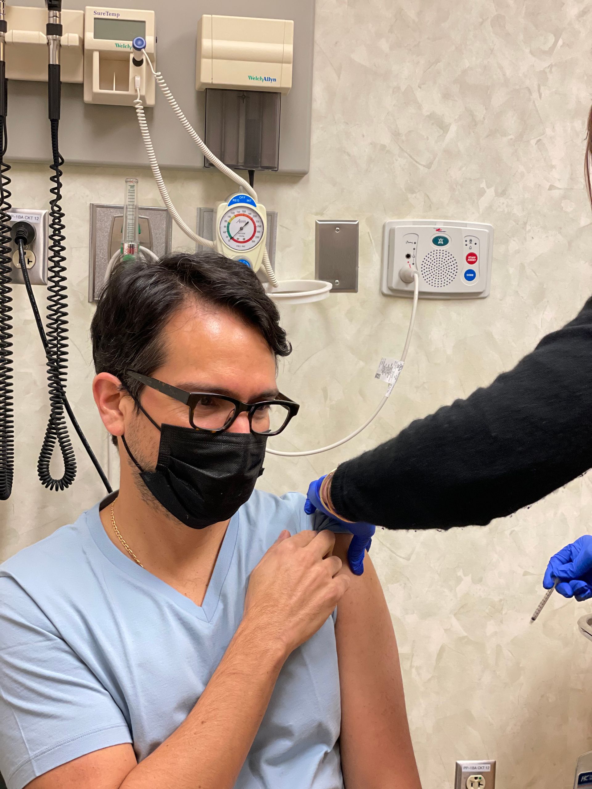 Man in a mask receiving a vaccine shot in his upper arm at a medical facility. A medical professional administers the injection.