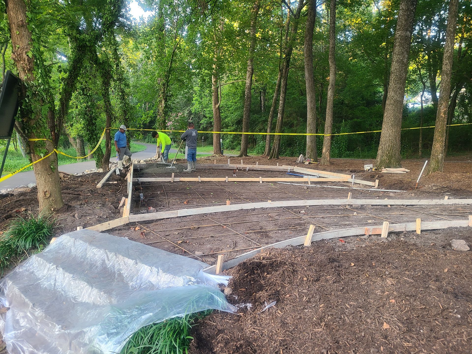 Construction site in a wooded area: workers laying concrete, wooden forms, caution tape, gray and brown tones.