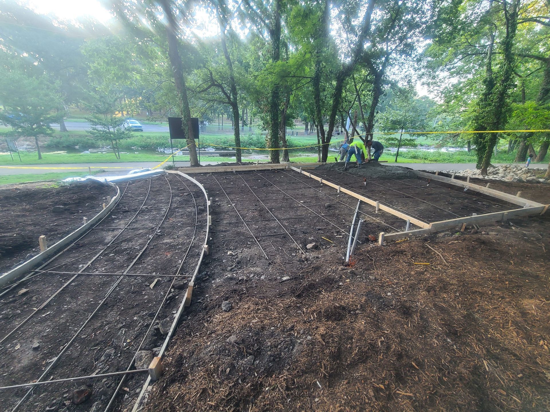 Construction site with dirt, metal reinforcing bars, and wooden frames. Workers in the background.