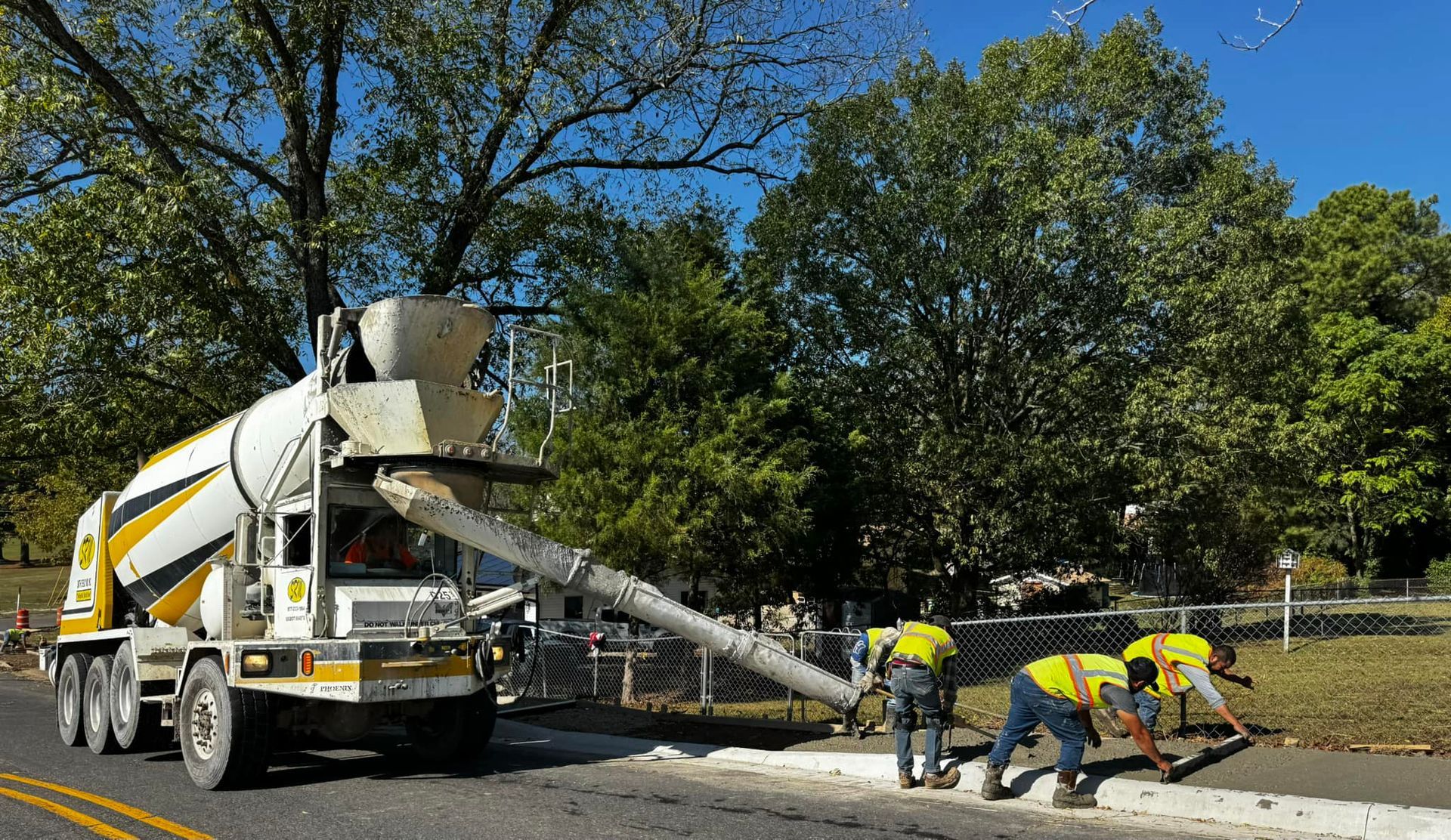 Cement truck pouring concrete; workers in vests finish edge along a road near trees.