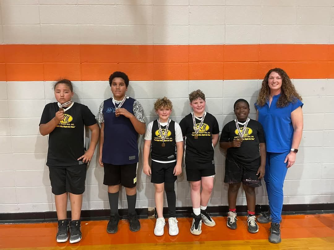 Group of young basketball players and a woman pose with medals against orange and white wall.