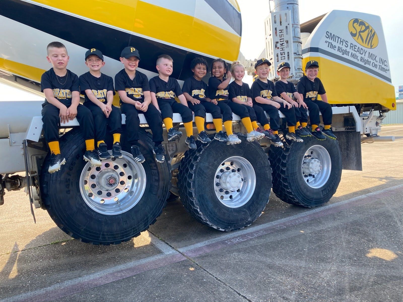 Little league baseball team sits on tires of a concrete truck. Black and yellow uniforms. Sunny outdoor setting.
