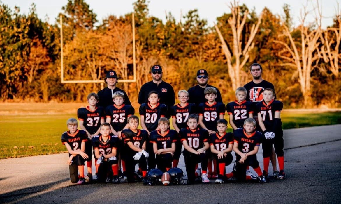 Youth football team posing on a field. Players in black and orange uniforms with coaches.