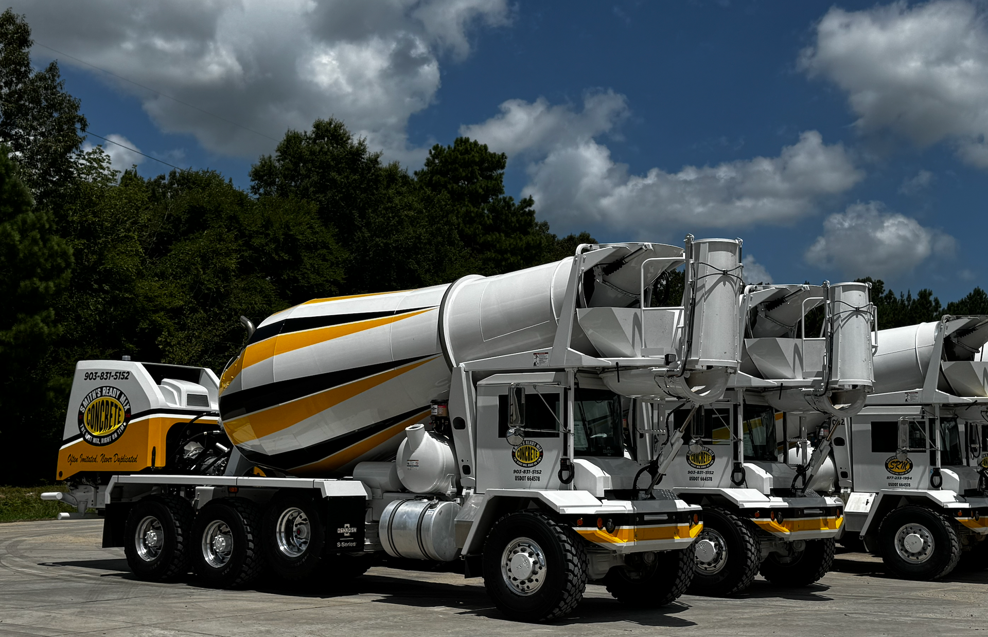 Cement trucks parked on a sunny day; white, yellow, and black design; trees in background.