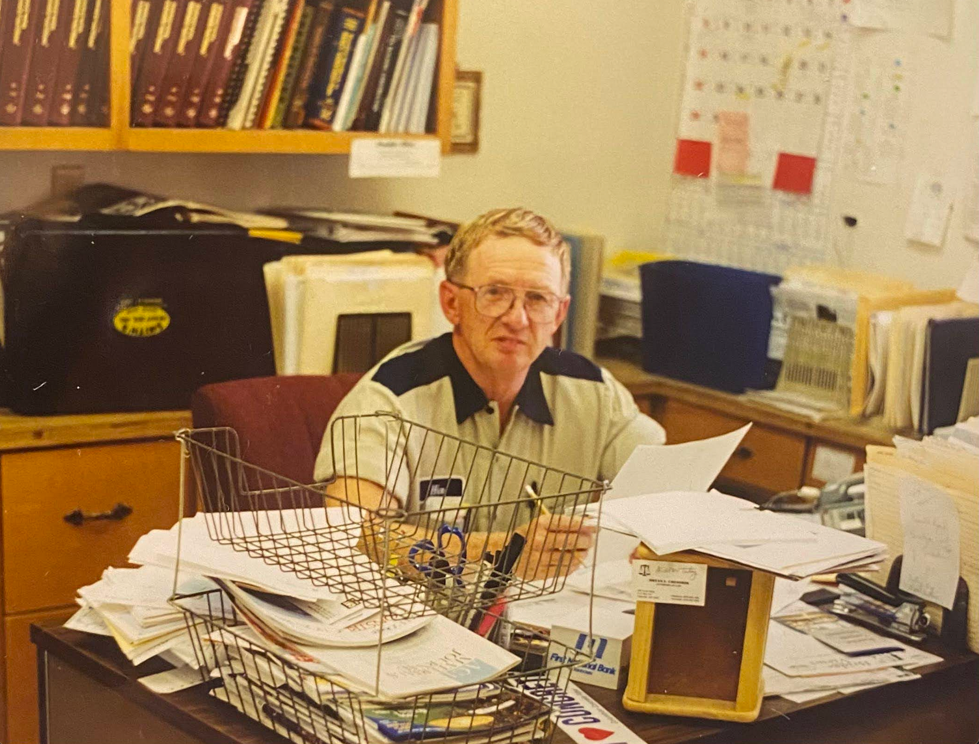 Man in glasses at a cluttered desk, holding papers. Bookshelves and calendar in background.