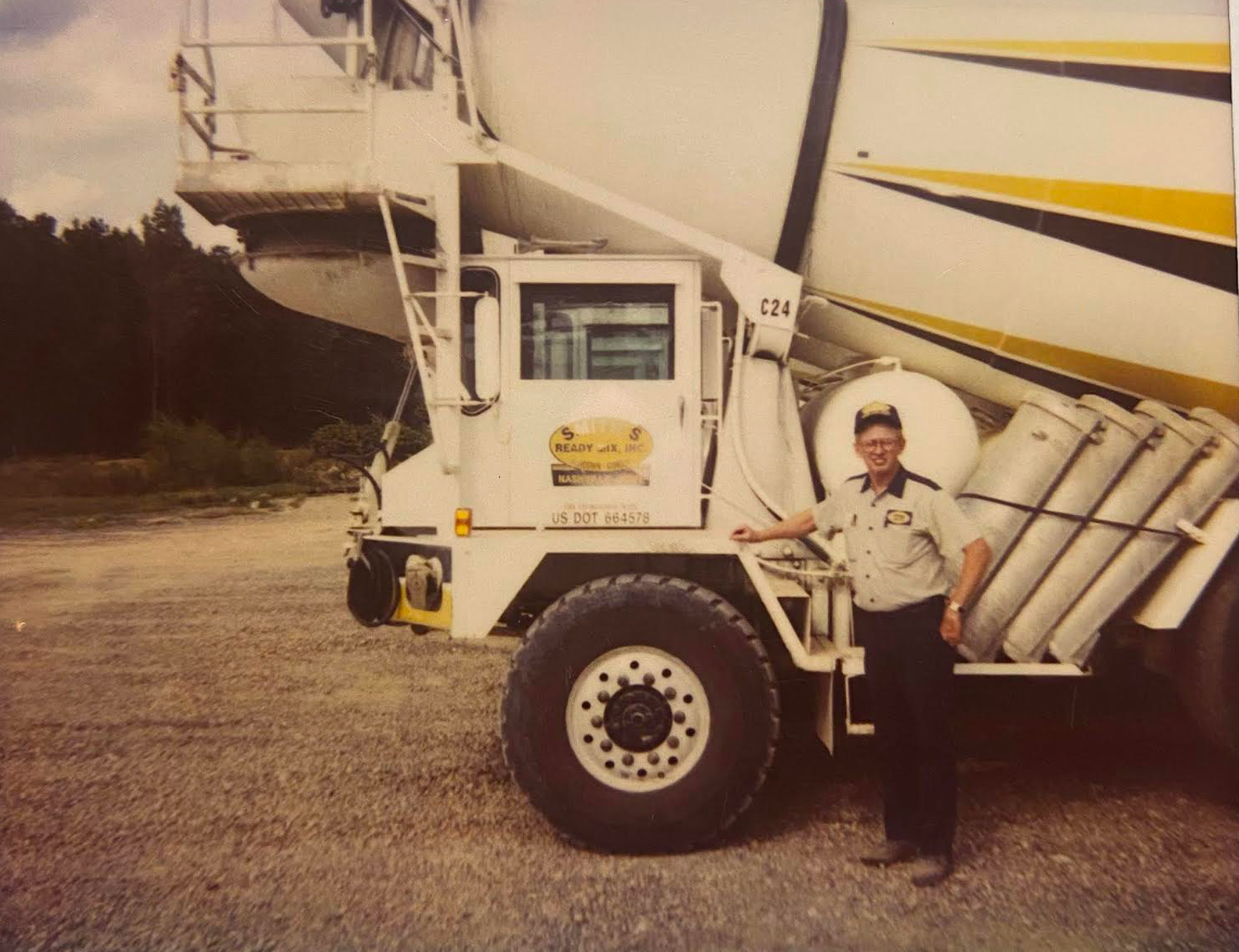 Man in uniform poses by a large white and yellow cement mixer truck. Gravel lot, overcast day.