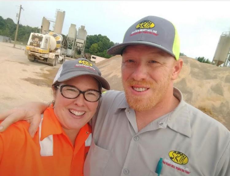 Two people smiling in construction gear, posing for a photo near equipment.
