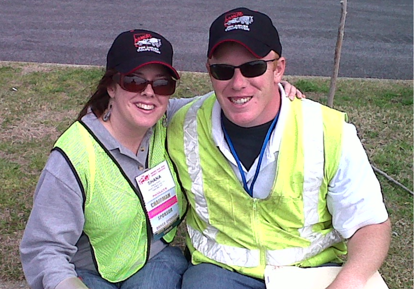 Two volunteers smiling, wearing reflective vests, sunglasses, and hats.