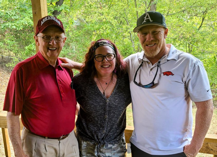 Three people smiling, posing outdoors: elderly man in red shirt, woman in tie-dye top, man with a hat in a white shirt.