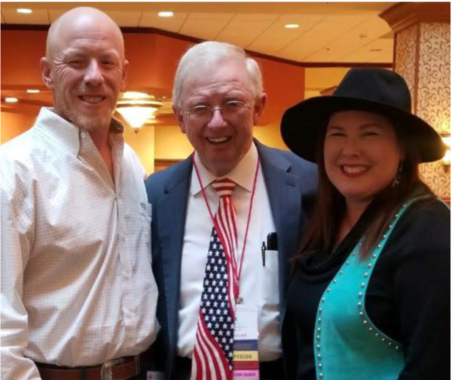 Three people smiling: a bald man, an older man with an American flag tie, and a woman wearing a hat. Indoors.