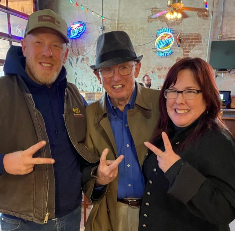 Three people smiling, making peace signs. Two men and one woman in a bar-like setting.