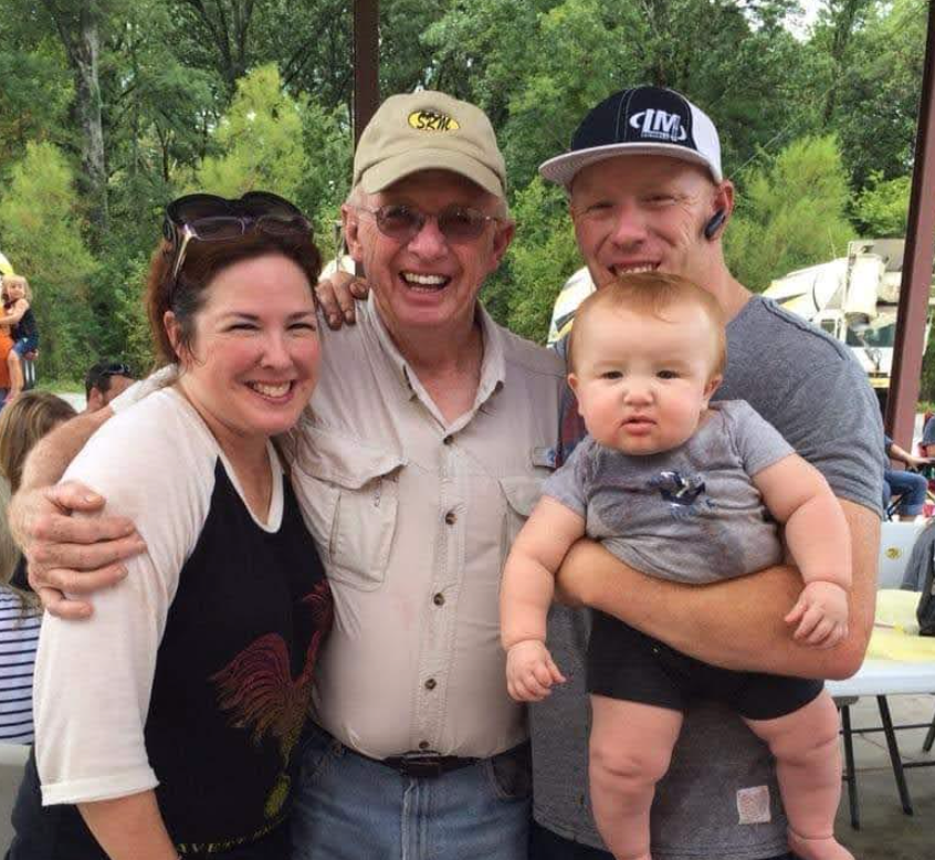 Four people smiling at the camera, including a baby held by a man, outdoors.