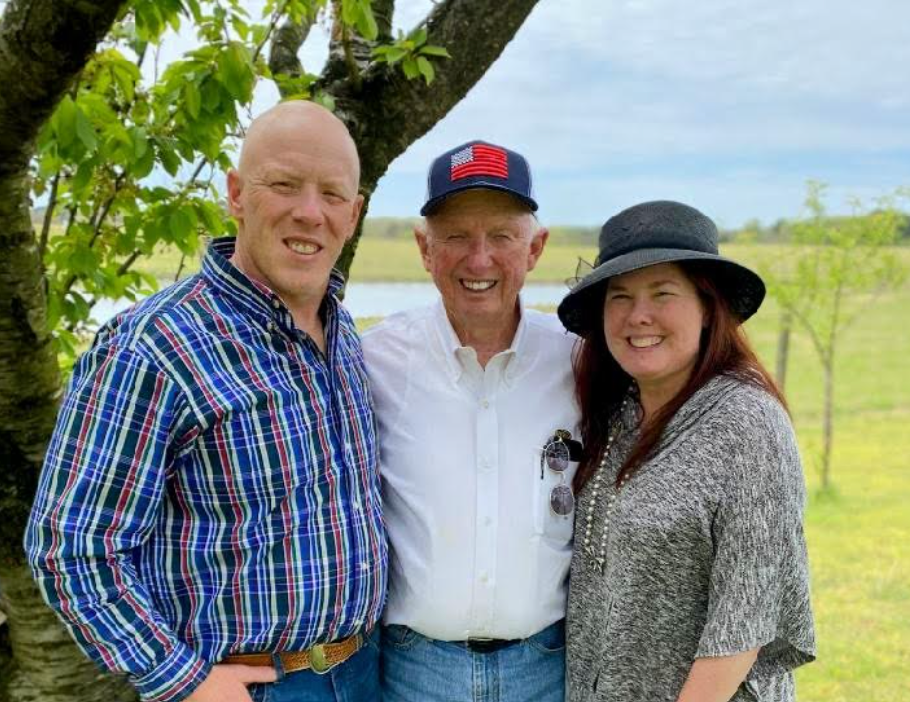 Three people smiling outdoors: a bald man, an elderly man in a cap, and a woman in a hat, with a pond and trees in the background.