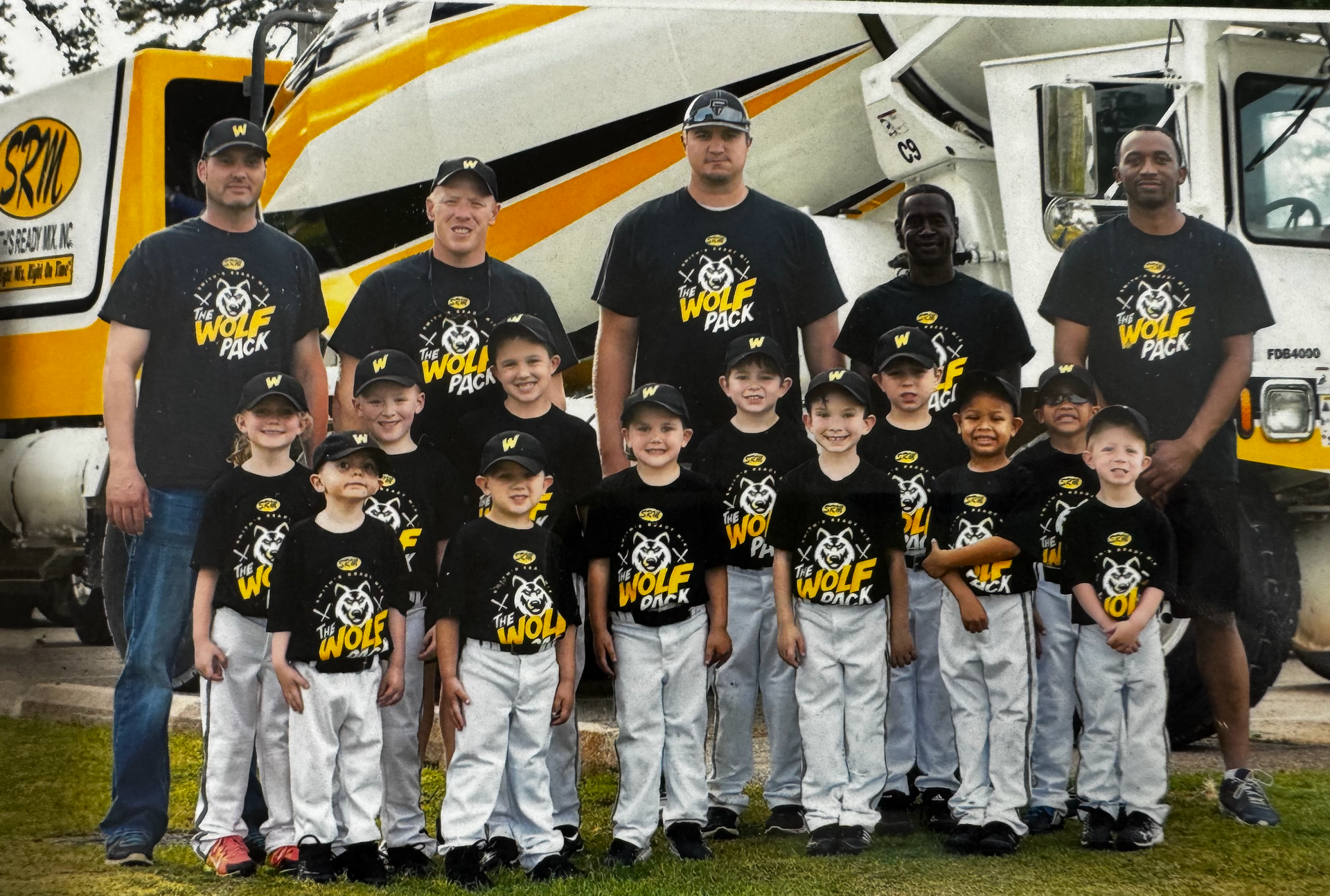 Youth baseball team in black and yellow uniforms posing with adult coaches in front of cement trucks.